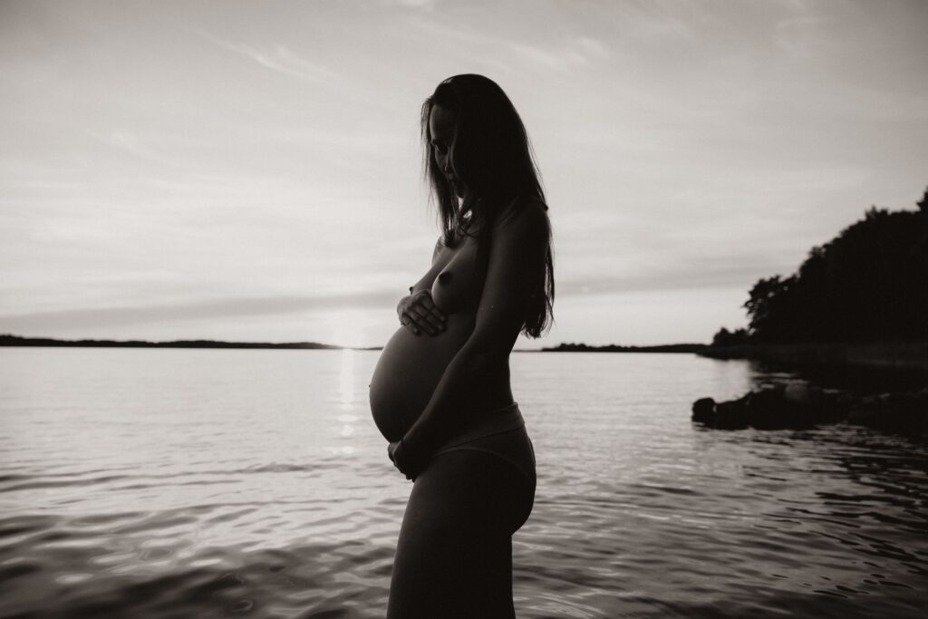 gravid kvinna står i vattnet i solnedgång och omfamnar sin mage