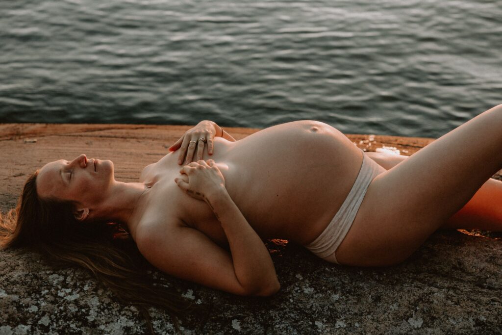 gravid kvinna med endast trosor ligger på en klippa vid vattnet och skyler sina bröst med sina händer