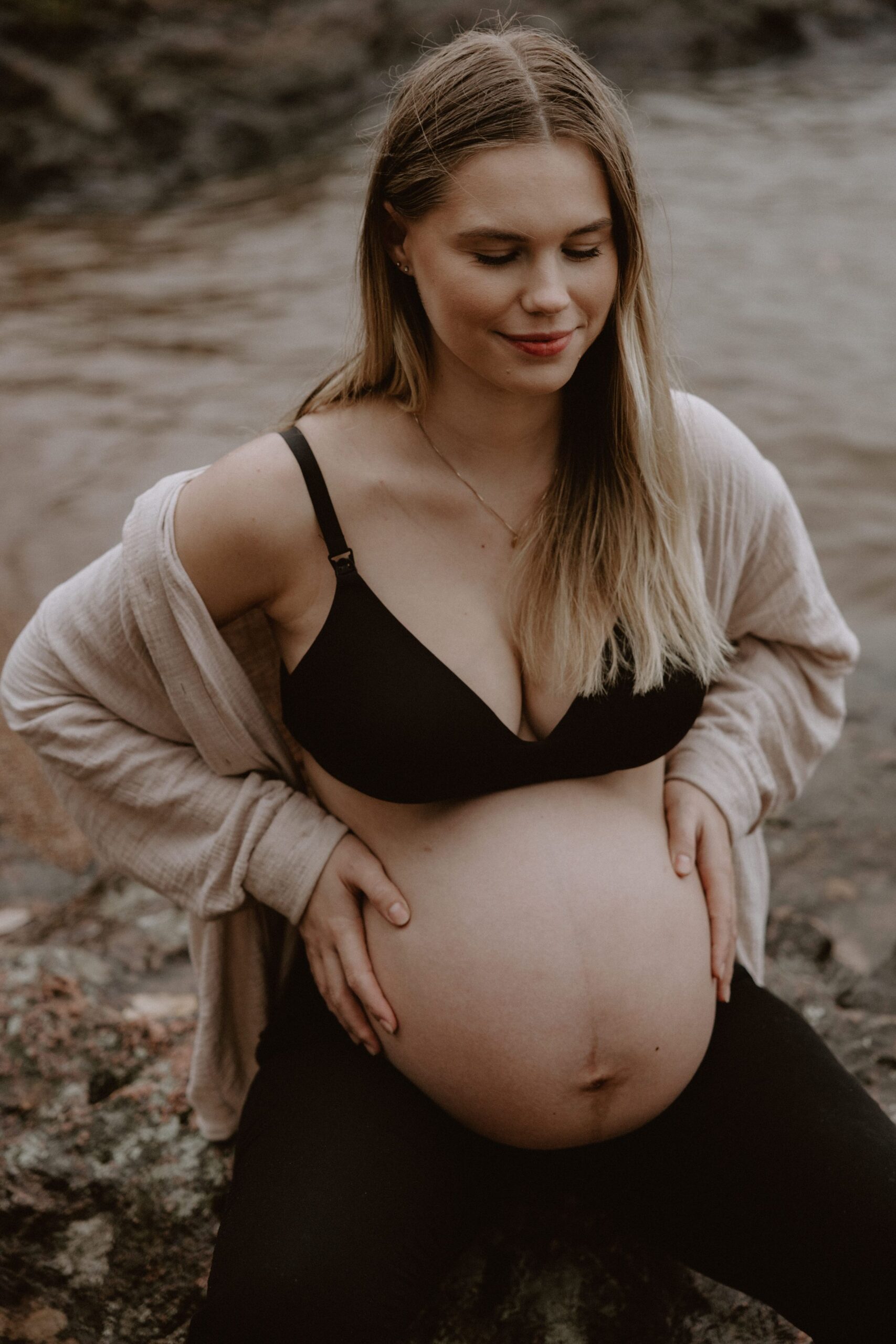 gravid kvinna sitter på en sten vid vattnet iklädd svarta byxor, bh och en beige skjorta