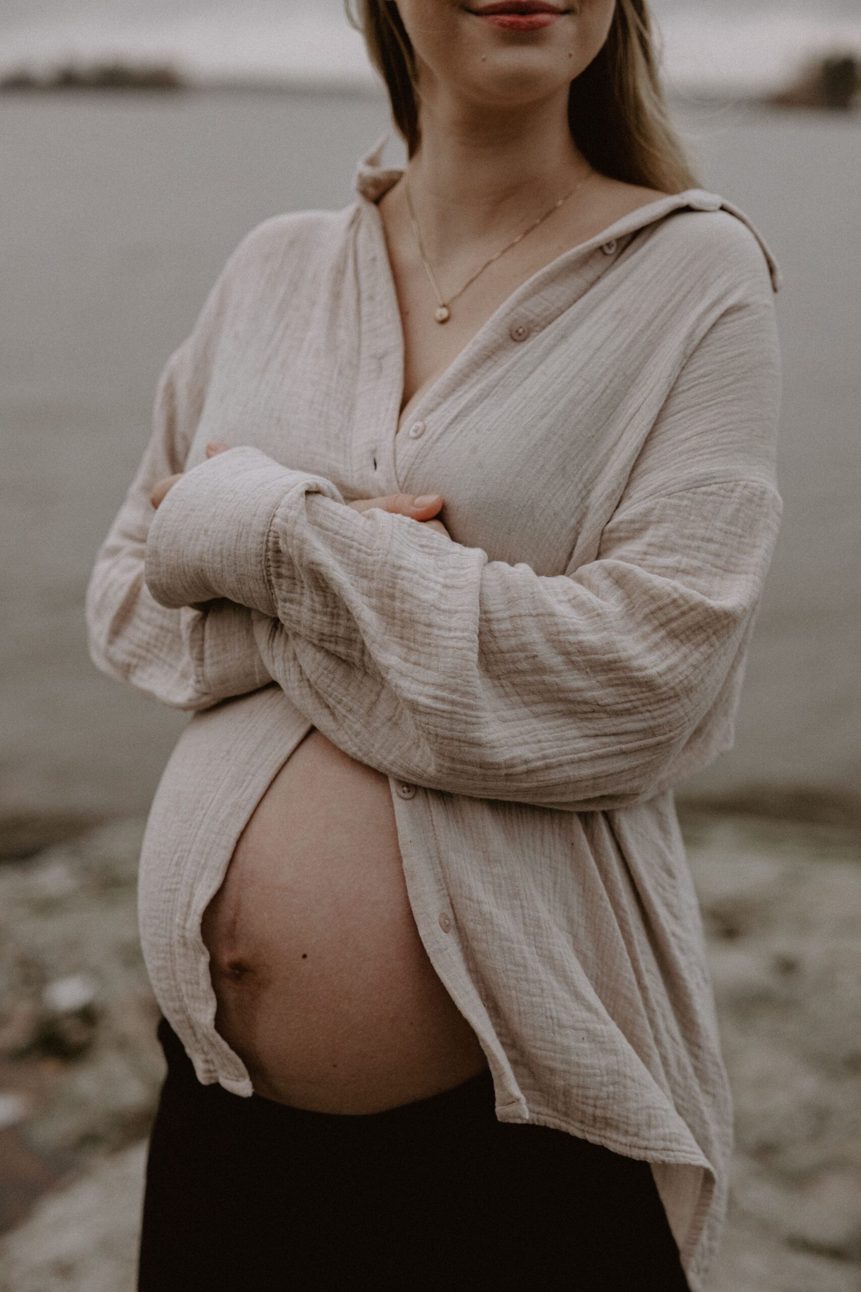 gravid kvinna iklädd en ljusbeige skjorta står på en klippa vid vattnet